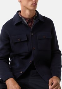 Granatowa kurtka z wełny w zestawie z dwiema kieszeniami na piersi, zapięciem na guziki i kołnierzem koszulowym; gładka tekstura i dopasowany krój.