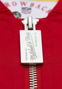 Roter Stoff mit einem silbernen Reißverschluss, versehen mit einem rechteckigen Etikett, das "Mitchell & Ness" und "Nostalgia Co. PHILADELPHIA, PA." eingraviert hat.