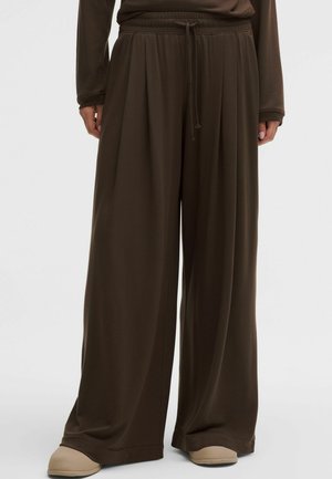 Weit geschnittene, braune Hose aus weichem Stoff, mit einem elastischen Bund und Kordelzug sowie feiner Textur, kombiniert mit hellem Schuhwerk.