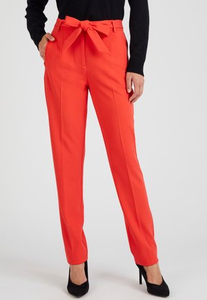 Rode op maat gemaakte pantalon met een strik rond de taille, voorzien van zijzakken en een gladde, gestructureerde stof. Gecombineerd met zwarte hoge hakken.