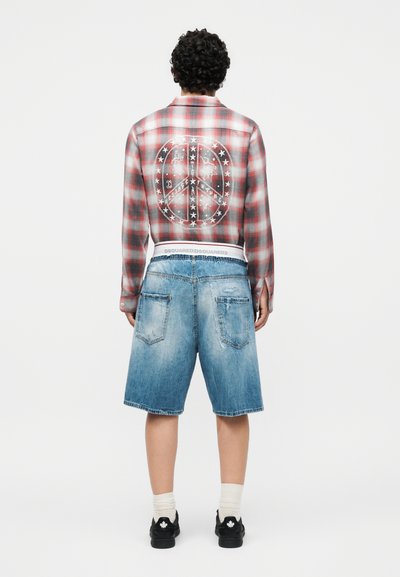 Άνδρας που φορά κόκκινο και γκρι πουκάμισο με καρό σχέδιο, μεγάλο λευκό σύμβολο της ειρήνης και αστέρια στην πλάτη, μπλε τζιν σορτς, λευκές κάλτσες και μαύρα αθλητικά παπούτσια.