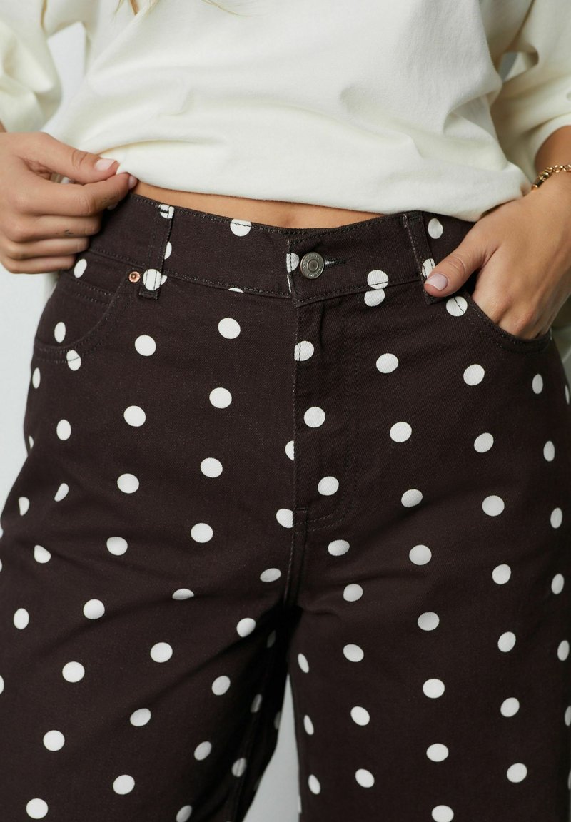 Vrouw met hoog uitgesneden donkere broek met witte stippen en een wit topje, handen die de tailleband verstellen en in de zak steken.