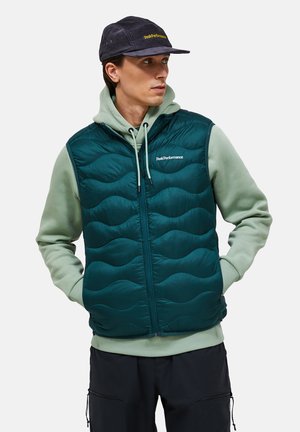 Uomo con un gilet trapuntato verde scuro sopra una felpa con cappuccio verde chiaro, pantaloni neri e un berretto nero, che guarda di lato con le mani in tasca.