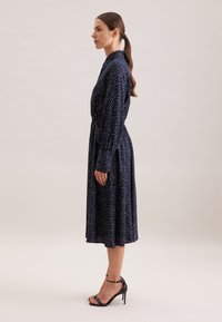 Abito blu navy realizzato in tessuto con pois bianchi, dotato di colletto, maniche lunghe e vita enfatizzata, abbinato a tacchi neri.