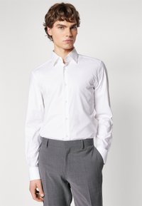 Camisetta bianca a maniche lunghe con un motivo testurizzato e colletto con bottoni, indossata con pantaloni grigi su misura. Presenta una vestibilità affusolata.