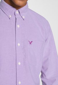 Camicia button-up di un leggero viola con colletto, dotata di bottoni bianchi e una tasca sul lato sinistro del petto. Include un piccolo logo di un'aquila viola sulla tasca.