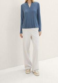 Sininen, pitkähihainen paita, jossa on V-pääntie ja rento istuvuus, yhdistetty valkoisiin, leveälahkeisiin housuihin. Asusteena on beiget tennarit.