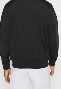 Svart idrottsjacka i slät tyg med texturerad yta. Har en ribbad midja och ärmslut. Bakifrån framhäver den en enkel design.