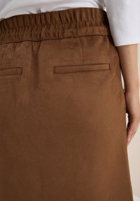 Jupe en daim marron avec une taille élastiquée, dotée de deux poches arrière minimales et d'une texture lisse.