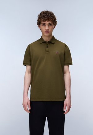 Jeune personne aux cheveux bouclés portant un polo vert olive à manches courtes avec un petit écusson logo et un pantalon noir sur un fond uni.