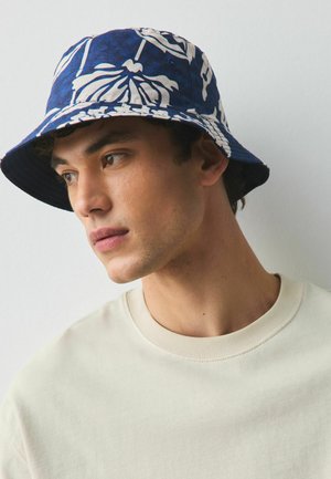 Mann trägt einen blauen Fischerhut mit weißen tropischen Blättern und ein hellbeiges Rundhals-Shirt und schaut zur Seite.