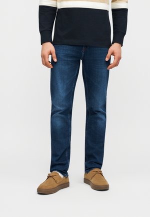 Άνδρας που φοράει σκούρο μπλε τζιν στενής γραμμής, μαύρο και μπεζ ριγωτό μακρυμάνικο πουκάμισο και καφέ σουέντ παπούτσια, στέκεται μπροστά σε απλό φόντο.