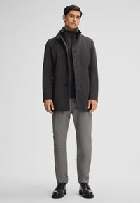 Cappotto nero con colletto strutturato e chiusura a bottoni, indossato sopra un dolcevita scuro. Pantaloni grigi a fantasia e scarpe nere stringate.