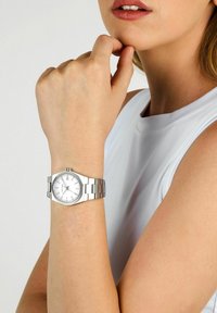 Orologio d'argento con quadrante bianco rotondo, design a bracciale con maglie e indici delle ore semplici. Indossato su un polso dalla carnagione chiara.