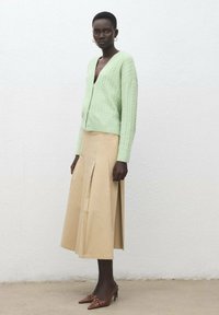 Svetlozelený sveter na zapínanie s vrkočovým vzorom, skombinovaný s béžovou midi sukňou s plisovaním, nosený s hnedými lodičkami s špicatou špičkou na textúrovanom pozadí.