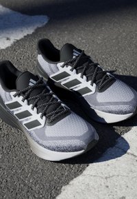 Baskets de sport grises et noires avec une tige en maille texturée, une semelle épaisse et un logo à trois bandes bien visible. Présente des lacets et des détails noirs.