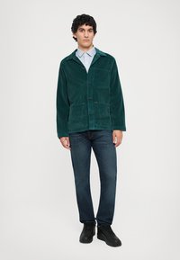 Green corduroy jacket with chest pocket, button front, worn over a light blue striped shirt, paired with dark jeans and black shoes.