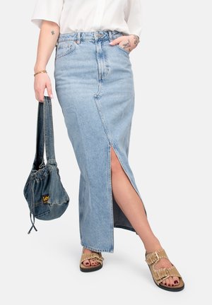 Maxi gonna di jeans blu chiaro con spacco frontale, vita alta e tasche laterali. Abbinata a una borsa di jeans blu scuro e sandali color nocciola.