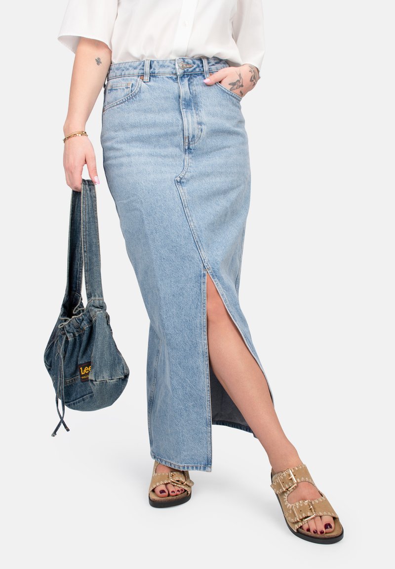 Lichtblauwe denim maxi rok met een voorkleedrelief, hoge taille en zijzakken. Gecombineerd met een donkergblauwe denim tas en tan sandalen.