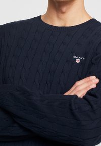 Personne portant un pull bleu marine à torsades avec le logo GANT sur la poitrine, bras croisés, montrant le motif tricoté texturé et le col rond côtelé.