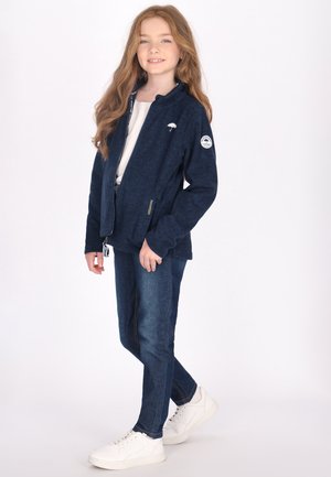 Mørkeblå zip-up jakke med struktureret finish, hvide accenter og logo patch; parret med mørke jeans og hvide sneakers.