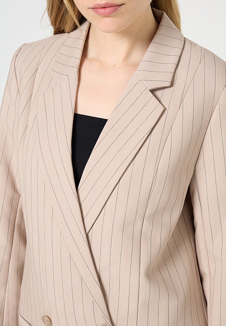 Femme portant un blazer beige à rayures sur un haut noir, se concentrant sur le haut du torse et le décolleté.