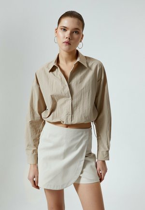 Beige kurz geschnittenes Hemd mit strukturiertem Stoff und langen Ärmeln, kombiniert mit einem hellbeigen Wickelrock. Beide Teile weisen minimalistische Designelemente auf.