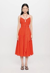 Vestido midi de algodão vermelho com detalhe em gota, cinto atado na cintura e saia ampla. Apresenta alças finas e um corte descontraído. Sandálias pretas com tiras.