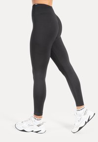 Leggings neri a vita alta con una texture liscia, caratterizzati da un design aderente e cuciture rinforzate, abbinati a scarpe da ginnastica bianche.