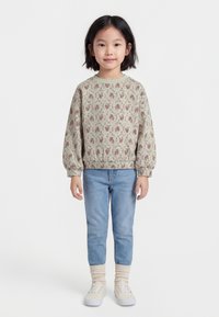 Jauna meitene stāv, ģērbusies ziedainā krekla ar garām piedurknēm, gaiši zilās džinsās, bēšās zeķēs un baltās sporta kurpēs, priekšplānā uz vienkārša fona.