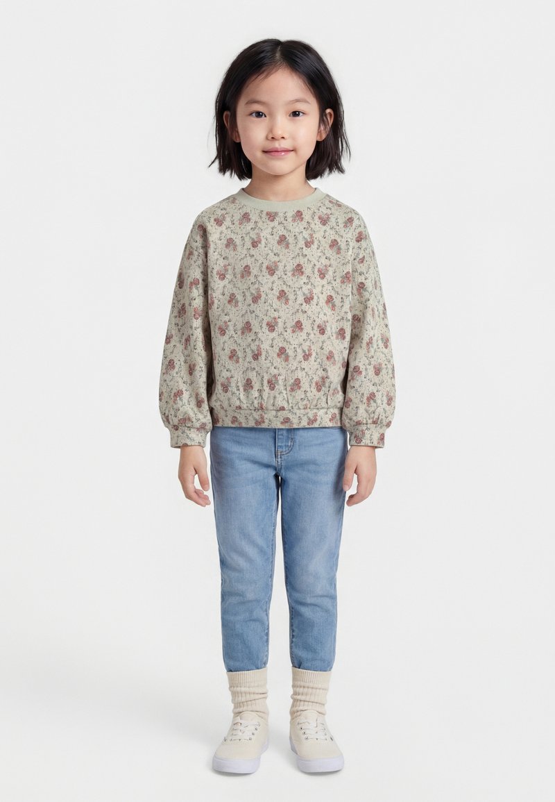 Jauna meitene stāv, ģērbusies ziedainā krekla ar garām piedurknēm, gaiši zilās džinsās, bēšās zeķēs un baltās sporta kurpēs, priekšplānā uz vienkārša fona.