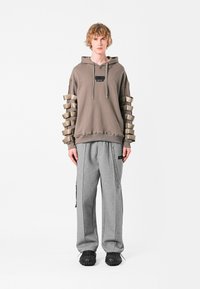 Sudadera marrón de gran tamaño con mangas de beige texturizadas y superpuestas, combinada con pantalones de chándal grises que cuentan con bolsillos laterales y un detalle de cadena.