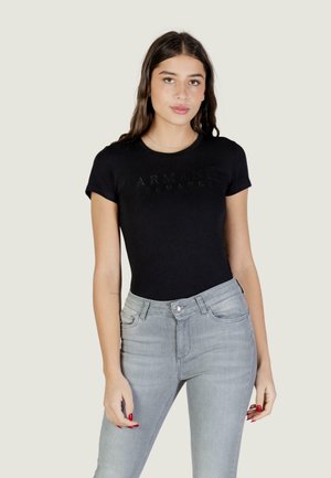 Giovane donna con i capelli lunghi castani indossa una t-shirt nera Armani Exchange infilata in jeans a vita alta grigio chiaro, con smalto per unghie rosso.