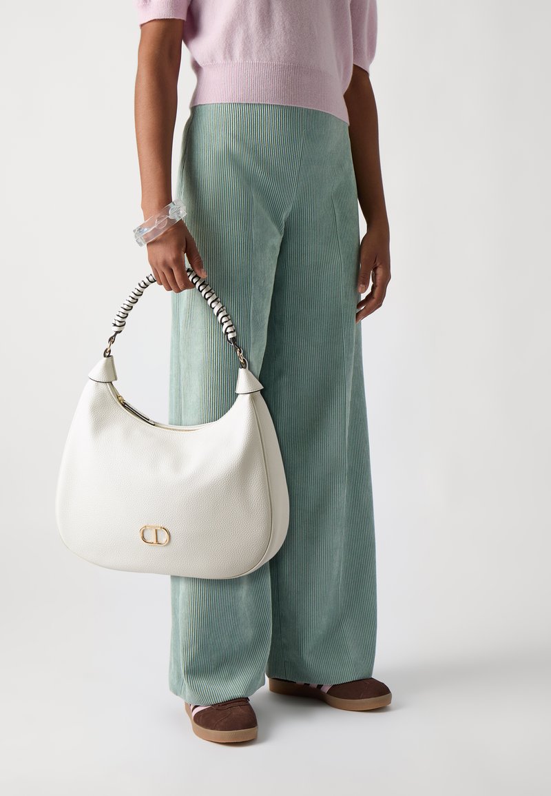 Borsa hobo bianca in pelle con manico a righe, dettaglio del logo dorato e forma soffice e arrotondata. Indossata con pantaloni in corduroy verde chiaro e scarpe marroni.