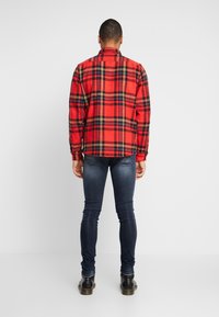 Röd och svart rutigt flanellskjorta, långärmad, med knäppning vid ärmsluten, rundad fåll, bärs med mörka slim-fit jeans och svarta stövlar. Bakvy.