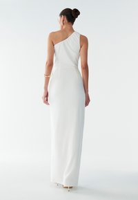 Vit aftonklänning med en axel, figurnära siluett, golvlång, slätt tyg och subtil detalj vid sidsömmen. Stilettklackar syns.