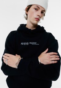 Svart hoodie med vit tryckt text, silverfärgad kedjebrassett och ringar. Bakgrunden är helt vit, vilket framhäver kläderna och accessoarerna.