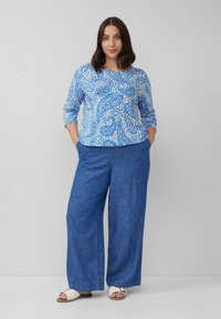 Bluse mit blauem Paisleymuster und Dreiviertelärmeln, kombiniert mit lockeren blauen Stoffhosen. Offen-toed Sandalen mit einem dekorativen Riemen.
