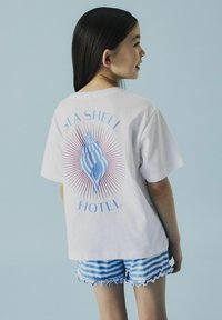 Biała koszulka z niebieską grafiką muszli i napisem "SEA SHELL HOTEL" z tyłu. Połączona z niebieskimi i białymi krótkimi spodniami w paski z falbankami.