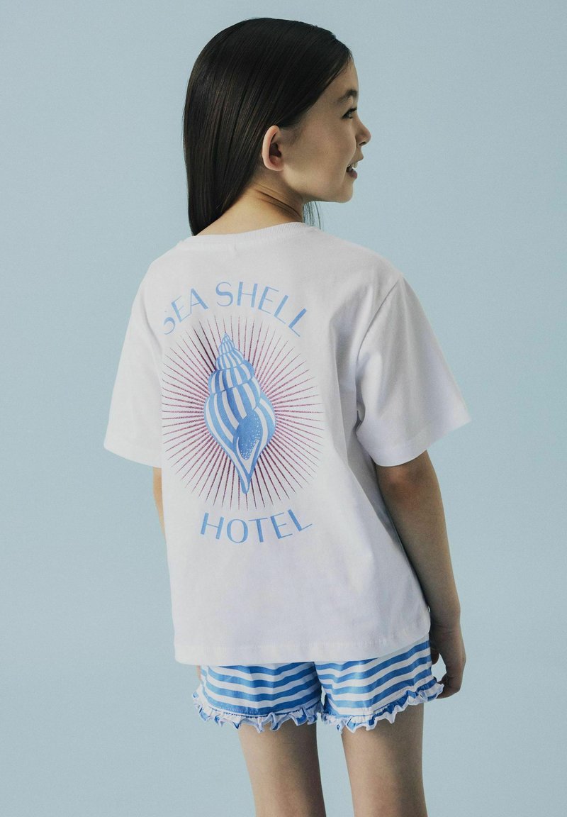 Biała koszulka z niebieską grafiką muszli i napisem "SEA SHELL HOTEL" z tyłu. Połączona z niebieskimi i białymi krótkimi spodniami w paski z falbankami.