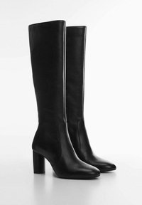 Mustat nahka polvipituiset saappaat, joissa on pyöreä kärki ja paksut korot, seisovat valkoisella pinnalla yksinkertaista vaaleaa taustaa vasten.