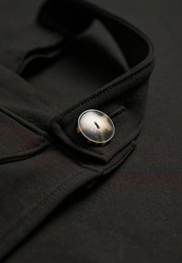 Nahaufnahme eines schwarzen Kleidungsstücks, das einen glänzenden Knopf am Kragen hervorhebt, wobei die Details der Textur des Stoffs und der Designelemente zur Geltung kommen.