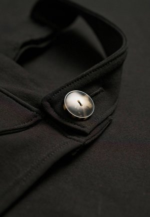 Close-up van een zwart kledingstuk met een glanzende knop op de kraag, waarbij de details van de stoftextuur en ontwerpelementen worden benadrukt.