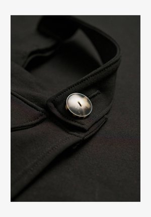 Nahaufnahme eines schwarzen Kleidungsstücks, das einen glänzenden Knopf am Kragen hervorhebt, wobei die Details der Textur des Stoffs und der Designelemente zur Geltung kommen.