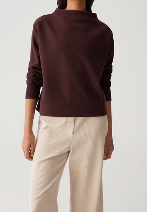 Femme portant un haut à manches longues marron foncé et un pantalon en velours côtelé beige ample, debout devant un fond clair uni.