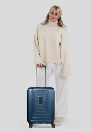Valigia rigida blu con un motivo a onde testurizzato, manico retrattile e quattro ruote, mostrata insieme a una persona con un maglione beige e pantaloni bianchi.