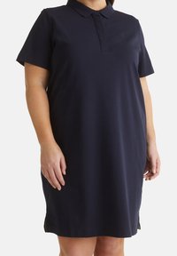 Femme portant une robe polo bleu marine à manches courtes avec col et patte de boutonnage cachée, debout sur un fond blanc.