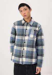 Button-up plaid shirt in blue, green, and cream with a chest pocket, quilted texture, and contrasting black buttons. Worn with dark pants.