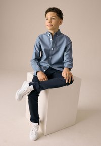 Camicia blu con bottoni, pantaloni blu navy e sneakers bianche. La camicia ha una finitura leggermente texturizzata e un piccolo logo sul petto.