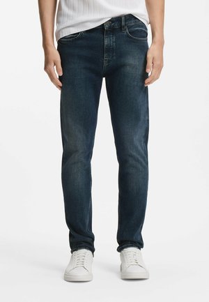 Hombre vistiendo jeans ajustados azul oscuro con detalles desgastados y zapatillas blancas, de pie contra un fondo blanco.